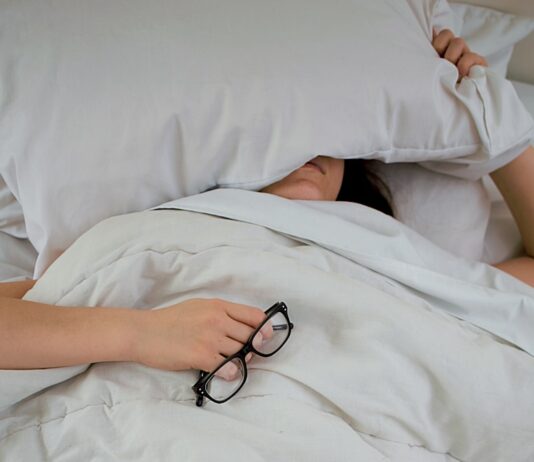 피로회복을 위한 에너지 충전 person lying on bed while covering face with pillow and holding eyeglasses